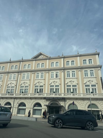 This image displays a street-level view of the Aveiro Palace building in Aveiro, Portugal, under a partially cloudy, light blue sky.
The Aveiro Palace is a multi-story building, approximately five floors tall, rendered in a light beige color with contrasting white decorative architectural elements. The facade features numerous white-framed, multi-paned windows. The two floors immediately above the ground level incorporate ornate, shell-like decorative motifs around the windows and a continuous balustraded balcony with several dark, spherical topiaries. A central pediment with an unidentifiable emblem and two decorative finials adorns the roofline. The building's name, "AVEIRO PALACE," is prominently displayed in dark lettering above the ground floor's central section.
At street level, the ground floor of the building presents a series of arched openings. Two arches on the left display "BPI" branding, indicating a bank branch or ATM. The central entrance to the Aveiro Palace is marked with an overhead "HOTEL" sign.
On the paved street in the foreground, two vehicles are visible. On the far left, the rear portion of a silver or light grey passenger car is partially captured, displaying a Portuguese license plate "15-01-RC". Centrally positioned on the street, a dark grey SUV with multi-spoke wheels is moving or parked, oriented towards the left.
On the sidewalk in front of the BPI section, a solitary individual dressed in dark clothing is observed walking to the left. Further to the right, near the hotel entrance, several additional indistinct figures are present on the sidewalk. Dark, short bollards line the curb between the street and the sidewalk.