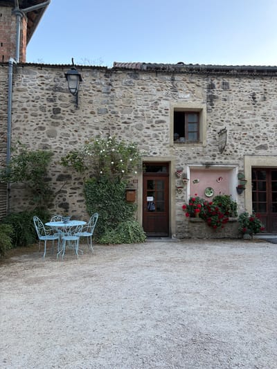 The image shows a charming courtyard scene in Saint Antoine l'Abbaye, France. A rustic stone building with wooden doors and windows dominates the background. A light blue wrought-iron table and four chairs are set in the gravel courtyard, suggesting an outdoor seating area. Lush green ivy and a white rose bush climb the stone wall, adding to the picturesque ambiance. Red geraniums overflow from planters, providing a vibrant splash of color. The sky above is a clear, pale blue. The overall impression is one of tranquility and rustic beauty.