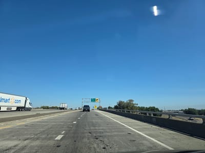 This media file depicts a view from a vehicle traveling on a highway. The scene is set in Sacramento, United States of America, under a clear blue sky, suggesting a sunny day. The highway has multiple lanes marked with white lines, and a concrete barrier runs along the right side.
In the foreground, the road surface shows some dark stains. In the middle ground, several vehicles are visible: a Walmart truck with "Walmart.com" branding is on the left, a dark-colored SUV is in the center lane, and other trucks and cars are further down the road. Overhead, a highway sign indicates "I-80 West San Francisco" and "20" with an arrow pointing to the right, suggesting an upcoming exit.
In the background, there are trees and some buildings, hinting at the transition from rural to urban or suburban landscape. The bright sunlight and clear sky indicate it is daytime, likely late morning or early afternoon. There is a bright, elongated light streak in the upper right portion of the sky, possibly a lens flare or reflection. The overall impression is one of normal highway travel on a pleasant day.