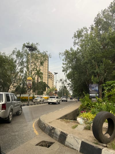 The image depicts a street scene in Falomo, Nigeria. The road curves gently and is lined with parked cars. A large, partially visible building stands in the background. Street signage and advertisements are present, including for MTN Fibre and a residential property named "Paramount Twin Towers," advertising "5-Star Living." A discarded tire sits on the curb. Lush greenery, including trees and bushes, borders the road. The sky is overcast.