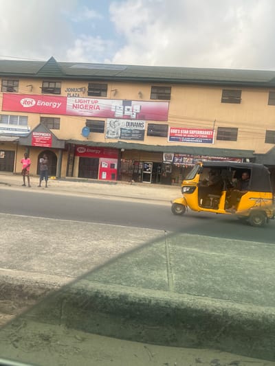 This image captures a daytime street scene in Elimgbu, Nigeria, featuring "JONUCK PLAZA" on Old Refinery Road, as identified by the "GOD'S STAR SUPERMARKET" signage. The two-story, tan-colored building has a dark green sloped roof equipped with visible solar panels. Multiple business signs are affixed to its facade, including a large red banner for "itel Energy LIGHT-UP NIGERIA," "BISHOPMEDICS LABORATORY & BLOOD TRANSFUSION," "DUNAMIS," and "GOD'S STAR SUPERMARKET." On the concrete sidewalk to the left, two males are standing; one wears a pink t-shirt and shorts, the other a striped t-shirt and dark bottoms. A yellow Keke Napep (motorized tricycle), containing at least two male occupants, is in motion on the paved road, traveling from right to left. The foreground shows a concrete curb and a section of the road, with a reflection indicating the photograph was taken from inside a vehicle. The sky above is partly cloudy.
