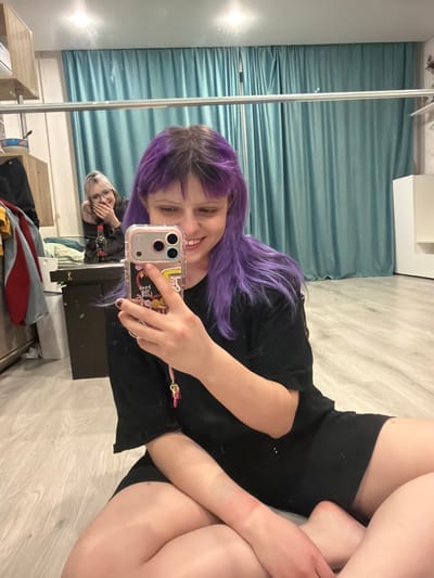 A mirror selfie features a smiling woman with vibrant purple hair and dark bangs. She's holding an iPhone with a patterned case. A reflection of another person is visible in the mirror behind her. The woman is wearing a black top and shorts, with her legs visible. The background includes light-colored curtains and a neutral-toned floor. Some furniture and clothing are also present in the reflected room.