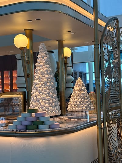 The image captures an indoor scene with two elaborate white meringue Christmas trees, suggesting a festive setting. The larger tree is positioned centrally, while a smaller one stands to its right. In the foreground, a stack of colorful, circular boxes (appearing in shades of green, lilac, and pale blue) is arranged in a pyramid on a marble-topped counter. The counter has a brass trim and is illuminated from below by warm lighting.
Behind the meringue trees, there are tall, golden columns and decorative light fixtures. The background reveals a sophisticated interior with built-in shelving units displaying various items, and large windows with a blueish tint. A decorative golden metal grate or screen is visible on the far right. The floor appears to be polished marble, reflecting the lighting and decor. No people are clearly visible, though a faint figure might be present in the reflection or background. The overall impression is of an upscale establishment, possibly a patisserie, hotel lobby, or a department store during the holiday season, situated in Arnavutköy, Turkey, a district known for its historic waterfront mansions and upscale atmosphere.