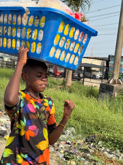 A young male, likely a boy, with dark skin and short hair, is depicted carrying a blue plastic laundry basket on his head, supported by his raised right hand. The basket contains numerous individually packaged items, predominantly yellow and white, appearing to be bread loaves or similar baked goods. He wears a vibrant, multi-colored floral-patterned t-shirt on a black background and a thin gold-colored bracelet on his left wrist, which is clenched into a fist. His eyes are partially closed, and his mouth slightly open, indicating concentration.
The outdoor scene, set in Jakande, Nigeria, is under bright daylight. The foreground and midground feature overgrown green grass extensively mixed with accumulated litter, including plastic bags and various debris. In the background, several vehicles (a red truck, a dark SUV, a white sedan) are parked near low-lying structures with metal roofs. A grey concrete pole, partially obscured but displaying "JOB" and a phone number ("0806 865837"), stands on the right. Palm trees and overhead power lines are visible against a clear blue sky.