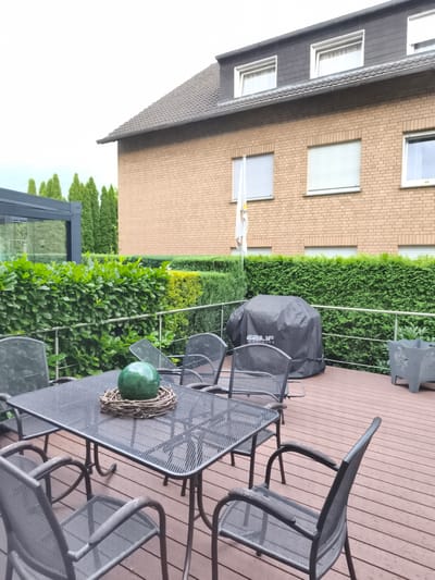 An outdoor seating area is depicted in Bad Rothenfelde, Germany. The scene is a wooden deck furnished with a metal dining table and four matching chairs. The table is adorned with a decorative green glass orb resting on a twig wreath. A black grill cover is placed on the deck behind the table, and a grey decorative planter with an embossed design is situated to the right. The background features a brick house with multiple windows and a dark tiled roof, partially obscured by lush green hedges and tall evergreen trees. The sky is overcast. There are no people visible in the image.