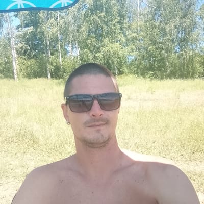 A selfie of a shirtless man wearing sunglasses is taken outdoors. He has short dark hair and a light beard. He is wearing a silver hoop earring in his left ear. The background shows a grassy field and trees under a blue sky. A portion of a blue and white striped umbrella is visible at the top of the image. The image is a photograph taken in the city of Panayot Hitovo, Bulgaria.