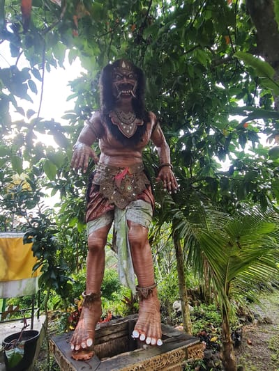A full-body, low-angle shot captures a life-size, demonic statue standing outdoors amidst lush green foliage. The statue, depicted in a dark tan, muscular form, is adorned with traditional Balinese attire. Its face is contorted in a fierce expression with prominent fangs and wide, staring eyes. The statue wears a crown, a beaded necklace, a ornate belt with a pink accent, and a sarong-like garment. Its feet, large and with white toenails, rest on a wooden platform with intricate carvings. The background is dominated by dense green leaves and branches of trees, with a bright, overcast sky peeking through. A yellow cloth is visible to the left, and a black planter with a plant is in the lower left corner. The ground appears wet, suggesting recent rain. The scene is located in Gianyar, Indonesia.