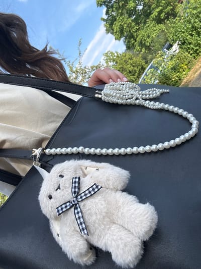 A close-up outdoor photograph shows a black handbag with a pearl necklace draped over it. A small, fluffy white bunny charm with a black and white checkered ribbon is attached to the bag. In the background, green trees and a bright blue sky with white clouds are visible. The image appears to be taken on a sunny day. The location is Bürgstadt, Germany.