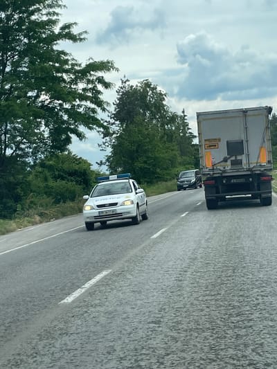 A daytime shot shows a white police car on an asphalt road lined by trees in Osenets, Bulgaria. A black car follows behind a trailer truck, all traveling in the same direction. The sky is cloudy. The police car has the word "POLICE" printed on it. The trailer truck has the license plate "A 5500 EM". The police car license plate is "PБ 5617 AY".