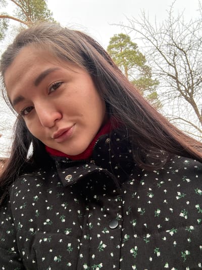 The image displays a close-up, low-angle selfie of a female individual outdoors in Kurovskoye, Russia. The woman has long, dark brown hair and brown eyes, looking directly at the camera with her head slightly tilted and lips subtly pursed. She is wearing a black jacket featuring a repeating pattern of small white flowers with green stems. A red, ribbed high-neck garment is visible beneath the jacket's collar. The jacket's collar has a visible silver snap button. The background consists of an overcast sky, with the upper sections of several trees visible. These include pine trees with green foliage and deciduous trees displaying bare branches, consistent with a cool season.
