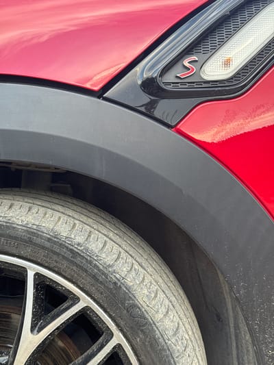 This media file is a close-up, eye-level shot of a red car's wheel well, focusing on the fender, tire, and part of the car's body. The car is red and has a black plastic fender flare. A red "S" emblem is visible on the black trim next to a turn signal light. The tire is dirty, indicating it has been driven on dusty roads. The rim of the wheel is black and silver with a multi-spoke design. The scene appears to be outdoors during the day, with natural light illuminating the car. The background is blurred, but it suggests an outdoor setting, possibly a road or parking area. There is no visible text in the immediate foreground, but some tire information is visible on the sidewall of the tire. The overall impression is a detailed, focused view of a part of a car, emphasizing its design and condition. The provided location context of Radanovići, Montenegro, suggests this is where the image was taken.