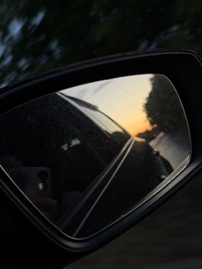 A side-view mirror reflects a sunset with trees and a car behind. The mirror is dirty and shows a reflection of a hand holding a phone. The car appears to be moving, as indicated by the blurred trees in the background. The time of day is likely evening, given the sunset. The location is Busmantsi, Bulgaria.
