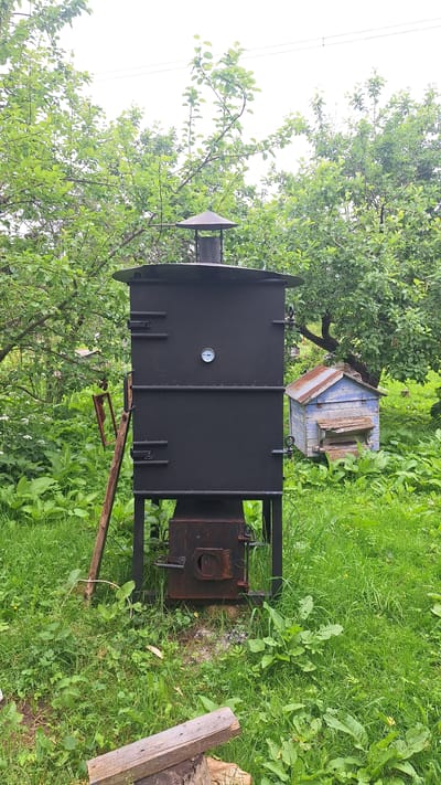 In a green, outdoor setting, likely in Kursieši, Latvia, a black, vertical, multi-chambered smoker stands prominently on metal legs. The smoker has two horizontal doors on its upper section and two similar doors on its lower section. A circular thermometer is affixed to the upper section. Beneath the smoker, a dark, rusty firebox with a circular opening is visible. To the right of the smoker, a small, weathered, light blue beehive with a rusty, corrugated metal roof is nestled amongst green foliage. In the foreground, a piece of weathered wood rests on a log. The background is filled with lush green trees and vegetation, suggesting a rural or garden environment.