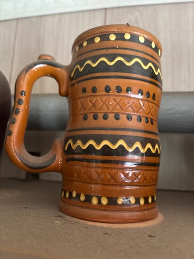 A close-up, eye-level, slightly low-angle shot captures a rustic, earthenware mug with a handle. The mug is decorated with horizontal bands of black and yellow wavy lines, yellow dots, and black dots. A crisscross pattern is incised into the orange-brown surface. The mug appears dusty, suggesting it is in a storage area. The background is out of focus, showing a shelf or surface. The overall impression is of an aged, possibly handmade item. The location of Ветлужский, Russia is not determinable from the visual information provided in the image.