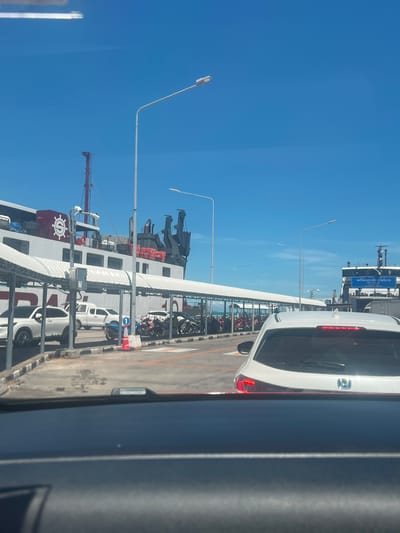 The image depicts a ferry terminal in Nathon, Thailand, on a clear, sunny day with a bright blue sky. In the foreground, the interior of a vehicle is visible, with the dashboard and windshield dominating the bottom portion of the frame. Through the windshield, a white car is seen from behind, stopped in what appears to be a queue.
Beyond the car, a covered waiting area with white pillars and roofing stretches along the terminal. Underneath this shelter, several motorcycles and a white pickup truck are parked. To the left of the covered area, a large white and red ferry is docked, its name partially visible. The ferry has a prominent circular logo on its side. In the background, another ferry, primarily black, is also visible, with Thai writing on a blue sign. Multiple lampposts are scattered throughout the scene. The ground appears to be paved, with painted lines marking parking spaces and a road. The overall impression is of a busy transport hub.
