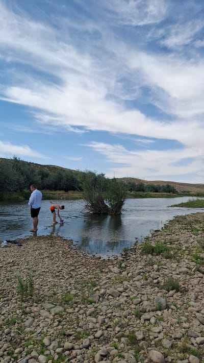The image depicts a river scene with two individuals. On the left, a man in a white shirt and black shorts stands in the shallow water near a gravelly bank. To his right, a boy wearing orange shorts is bent over in the water, holding a purple bucket. The river has a visible current and is lined by a rocky shore with sparse vegetation. Across the river, a treeline of green foliage stretches along rolling hills under a bright blue sky with wispy white clouds. The location is identified as the river in Boraldai, Kazakhstan.