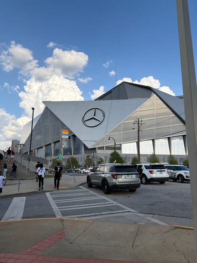 The image shows the exterior of Mercedes-Benz Stadium in Atlanta, Georgia. The stadium's distinctive architecture with the large Mercedes-Benz logo is prominent. Several vehicles, including a MARTA police car, are parked along the street adjacent to the stadium. Pedestrians are walking on the sidewalk and stairs leading up to the stadium entrance. The sky is blue with white clouds. A Marta transit sign is visible.