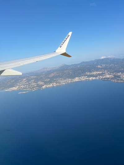 An aerial photograph depicts a coastal landscape observed from a Ryanair aircraft. The white wing of the aircraft, bearing the "RYANAIR" logo in blue text on its wingtip, occupies the upper left portion of the frame. Below the wing, a vast body of deep blue water, identified as the sea, fills the lower half of the image. Along the horizon, a densely populated coastline is visible, featuring numerous light-colored buildings and structures situated amidst green vegetation. This coastal area and its mountainous backdrop are consistent with the region of Sol de Mallorca, Spain. The sky above is clear and intensely blue, suggesting bright daylight conditions. No individuals, explicit actions, or interactions are discernible.