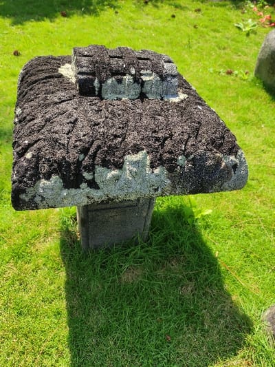 The scene depicts a heavily weathered, dark grey stone monument or sculpture situated on a vibrant green lawn in Staufenberg, Germany. The central object consists of a broad, roughly rectangular stone slab resting on a squarish, smaller stone base. The top surface of the slab is extensively covered with a thick, dark, textured organic growth, appearing black with intermittent patches of lighter grey stone visible, particularly along the perimeter and where the growth is less dense. Along the central axis of the top slab, several distinct, raised, rectangular block-like features are visible, also partially obscured by the dark growth. The supporting base exhibits similar weathering to the exposed stone of the slab. The ground is covered by uniformly green, well-maintained grass. Strong, direct sunlight illuminates the scene from the upper right, casting a prominent, dark shadow of the stone structure onto the grass, extending towards the lower left. In the mid-ground and background, additional green grass is visible, along with scattered small, dark brown objects, possibly pinecones. A portion of a larger, rounded grey stone is visible on the far right edge of the frame. No individuals, animals, or active interactions are present.