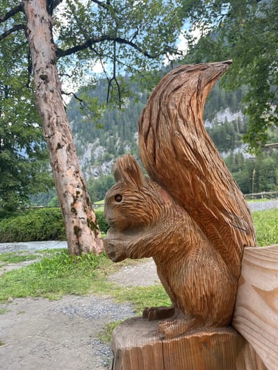 This is a vertical, close-up, eye-level shot of a wooden squirrel sculpture located in Wengen, Switzerland. The sculpture is positioned on the right side of the frame, carved from a light brown wood with visible grain and chisel marks, depicting a squirrel with its bushy tail curled over its back. The squirrel's head is turned to the left, with carved eyes and a mouth. The sculpture is mounted on a wooden base, which extends to the lower right corner of the image.
To the left of the sculpture is a large, textured tree trunk with peeling bark, its branches extending upwards and providing shade. In the background, a river flows to the left, bordered by lush green grass and vegetation. Beyond the river and vegetation, a steep, rocky mountain face covered with dark green trees rises to the sky. The sky above is partly cloudy with patches of blue. The overall environment suggests a natural, possibly park-like setting in the Swiss Alps. No people are visible in the image.