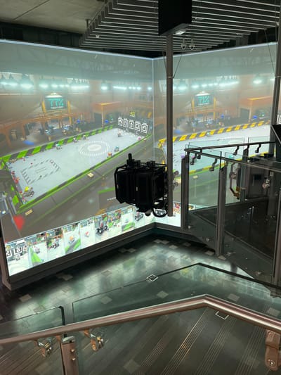 This rotated image, taken in an indoor setting likely in Brisbane City, Australia, captures a dynamic interactive floor display viewed from an elevated perspective. The central subject is a large, brightly lit projection on the floor, showcasing a virtual racing game or interactive experience. The game features a track with a prominent circular central area, surrounded by a workshop-like environment populated with numerous virtual vehicles resembling go-karts or small cars, some appearing to be modular or block-built.

Visible text and graphics on the display include "TODAY'S BEST," "72%," and "41%," alongside several icons that likely represent game scores or achievements. Faintly visible, upside-down text near the bottom edge of the projection reads "IN AUSTRALIA."

Above the floor display, a dark, multi-component piece of equipment, possibly a projector system or motion-tracking cameras, is suspended from an overhead structure, indicating the interactive nature of the setup. The immediate foreground features a sleek glass barrier and a polished metal handrail, suggesting the vantage point is from a mezzanine or an upper level overlooking the main area. The floor beneath the glass barrier is tiled in a checkered pattern of black and grey squares. The overall scene suggests a modern entertainment, gaming, or educational venue. No people are directly visible in the image.