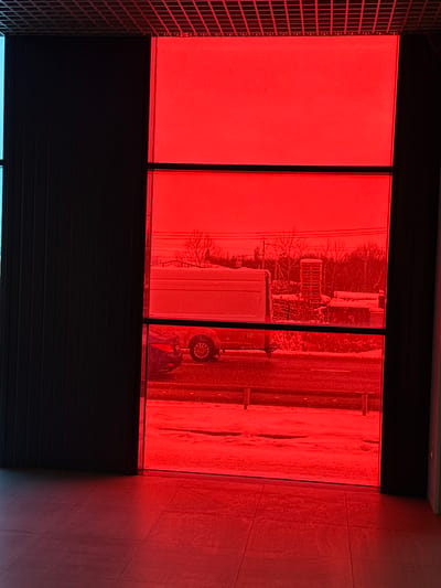 The image depicts an interior perspective looking outward through a large, multi-paned window with a prominent red tint, overlooking an exterior scene in Druzhny, Russia. The interior foreground features a light-colored, tiled floor reflecting the red hue of the window. A grid-patterned ceiling is visible at the top, and dark vertical panels or blinds are present on the left, partially obscuring a narrow, faintly blue-tinted window section.
The exterior scene, viewed through the red tint, indicates cold weather with snow covering the ground. A cleared, dark road traverses horizontally across the mid-ground, flanked by a snow-covered verge and small, dark barriers. A light-colored panel van is positioned on the right side of the road, facing left, its side visible. Behind it and slightly to the left, the rear section of a dark-colored sedan is partially visible. In the background, beyond the road and snow, bare deciduous trees and utility poles with power lines are present. A light-colored, possibly cylindrical stack of objects or an industrial structure is discernible in the far right background. No people are visible in the scene.