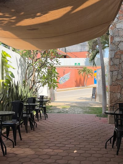 This outdoor scene in Brisas de Zicatela, Mexico, features a brick patio area with multiple black chairs and tables. A light brown canopy partially obscures the left side of the frame. Through the gap between buildings, a streetscape is visible, displaying colorful buildings, including an orange one with a wing mural. A palm tree stands nearby. Vegetation is present along the bottom edge and between the patio and the street. The environment suggests an outdoor dining or seating area of a cafe or restaurant.