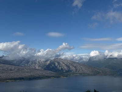 The image depicts a vast, serene landscape of mountains and a body of water, likely a fjord, under a bright blue sky dotted with scattered white clouds. The foreground is dominated by the dark blue water, reflecting the sky. Beyond the water, a textured, rocky mountainside rises, its surface appearing weathered and uneven. Further in the distance, more rugged peaks emerge, some partially obscured by white, puffy clouds that are clinging to their slopes. A strip of green forest separates the rocky terrain from the water's edge, with a few scattered structures suggesting a small settlement. The overall impression is one of natural grandeur and tranquility. The geographical location is identified as Flostrand, Norway.