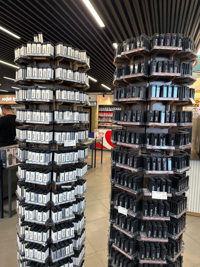 The image shows shelves of personalized lighters with names on them. They are arranged in rows. The lighters are black and have labels with white writing. In the bottom-left corner, a person is partially visible, wearing a dark shirt and looking down, likely browsing the products. The background includes store shelves with various items, and ceiling details. The text "soğuk İç" is visible, indicating a food and beverage section. Based on the OCR text mentioning "ALGI," which could refer to the Algida brand (ice cream), there are Algida freezers in the background. The text "ALMAK YAS" implies a restriction, and "ALIMI DISE" seems to be a sales-related message. It is located in a store, presumably in the city of Gaziosmanpaşa, Turkey.