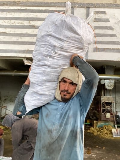 In Shurandam, Afghanistan, a man carries a large, white sack over his right shoulder. He wears a light blue, button-down shirt with sleeves rolled up to the elbows, and a beige head covering. The shirt and his face are visibly dirty. His arms are raised to support the sack. The setting appears to be an industrial space. A person is partially visible in the lower left corner, bending over. Equipment, including what seems to be a large machine and a shovel, is visible in the background. The walls are pale green and there is a horizontal band of gray and white. The environment suggests labor-intensive work.