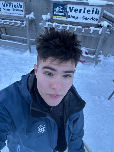 A young male with short, spiky dark hair and brown eyes looks directly into the camera, taking a selfie outdoors in a snowy environment. He wears a dark blue waterproof jacket with a white circular logo featuring a stylized turtle or similar animal on the left chest, over a black zippered fleece. His mouth is slightly open. Behind him, the ground is covered in deep snow, with visible tracks. A wooden outdoor rack structure, also partially covered in snow, stands against a light-colored wall. Two signs are visible on the wall behind the structure: the left sign is partially obscured, displaying "Verleih Shop - Service" and "www.angertal.com"; the right sign, mostly visible, reads "TOP-RENT," "ANGERTA L," "GASTEIN," and "Verleih Shop - Service." The scene is consistent with a winter ski resort location, specifically Angertal in Gastein, Austria.