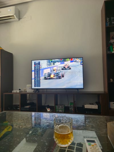An indoor scene within a residential property located in Picassent, Spain, depicts a living area from a low vantage point looking towards a television and surrounding furniture.

In the immediate foreground, a reflective glass coffee table holds a half-full glass of pale amber liquid, consistent with beer, topped with a head of foam. To its right, a white remote control with green, orange, yellow, and white buttons rests on a folded white document or receipt. Partially visible to the left on the table are blurred yellow and dark objects.

A large, flat-screen television is centered on a dark brown, multi-compartment media console. The television prominently displays a Formula 1 race, showing multiple cars on a track with safety barriers. The on-screen overlay indicates "LAP 76/78" and lists drivers, with "VER" (Verstappen) identified as the leader with "OUT" status listed next to his name and several others, suggesting a particular moment in a race broadcast or replay. A Red Bull racing car is clearly visible on the track.

On the left side of the media console, a white DIGI branded modem/router stands upright next to a smaller white network device, both accompanied by visible cabling. The central compartments house a black Xbox gaming console, a power strip with an illuminated red power button, and multiple video game cases with green spines (identifiable titles include "Halo," "Call of Duty: Black Ops," and "Call of Duty: Ghosts"). To the right, more game cases, a white packaged box, and additional electronic devices are stored within the console's shelves.

The wall above the console is a textured, light grey color. To the upper left, a white Haier split-system air conditioning unit is mounted high on the wall, displaying "25" on its digital screen, indicating a temperature setting. A white conduit runs horizontally from the right side of the unit. To the far right of the scene, a tall, dark brown multi-shelf bookshelf contains various books and decorative items, including what appear to be "Jack Daniel's" branded boxes, a small flag with red and green stripes, and a small white figurine. No individuals are visible in the frame.