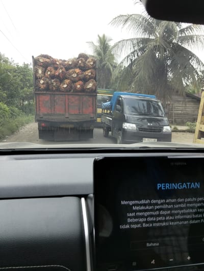 This is a photo taken from inside a vehicle in Awegeutahpaya, Indonesia. The vehicle is on a dirt road with some vegetation on the left side. In front of the vehicle, there is a red truck loaded with palm oil fruit. To the right of the red truck, there is a black pickup truck with a blue tarp covering its bed. The pickup truck has a license plate that reads "BK 8022 NF". In the background, there are palm trees and wooden structures. The foreground of the image shows the dashboard of the vehicle the photo was taken from, with a car infotainment screen displaying a warning message in Indonesian. The screen shows the Toyota logo at the top.