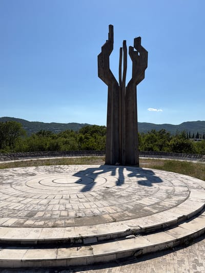 The image captures a low-angle perspective of a modernist monument against a backdrop of a clear blue sky and a distant hillside. The monument, constructed of dark, weathered concrete or stone, consists of a tall, abstract central element with angular, wing-like extensions at the top. The monument casts a distinct shadow on a circular, multi-tiered platform made of light-colored paving stones. The surrounding environment includes a green, vegetated area between the monument and the distant hillside. This is the memorial complex of Barutana near Podgorica in Montenegro.