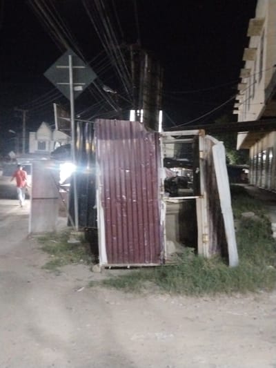 The image depicts an outdoor scene at night, possibly in a village or town setting in Keudegeudong, Indonesia. The foreground is dominated by a rough, dirt or gravel path. To the right, there is a pile of corrugated metal sheeting, some of which is reddish-brown and appears to be a makeshift barrier or roofing material. Adjacent to this, a white sheet of corrugated material leans against a metal frame.
In the middle ground, a person wearing an orange shirt and light-colored pants is walking away from the camera, towards the left side of the image. They appear to be on the dirt path. To the left of the person, there is a tall, light-colored structure, possibly a building or a large signpost, with a diamond-shaped sign attached to it. This sign has a white cross symbol on a dark background. Several utility poles and a dense network of overhead wires are visible, suggesting an urban or semi-urban environment with infrastructure.
In the background, there are glimpses of buildings. On the right, a multi-story building with a light-colored facade and visible windows or openings stretches across the frame. On the left, a lighter-colored building with a pitched roof is partially visible. The overall scene is dimly lit by artificial light sources, and the sky is dark, indicating it is nighttime. The presence of overgrown grass in some areas suggests a somewhat unkempt or developing environment. There is no visible text in the image. The atmosphere appears to be mundane, possibly a street scene or an alleyway at night.
