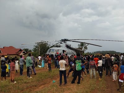The image displays a grey military-style transport helicopter landed on a reddish-brown dirt field in the city of Beni, Democratic Republic of the Congo. A large crowd, consisting of hundreds of individuals including adults and children, is gathered around the helicopter, primarily observing it. The helicopter's main rotor blades are extended, and an open door is visible on its side, near where people are standing. The ground is characterized by dry, reddish-brown earth with patches of sparse green vegetation. In the background, two buildings with red-tiled roofs are visible to the left, and various green trees and bushes, including a palm tree, extend towards a distant tree line. The sky above is uniformly overcast with grey clouds.