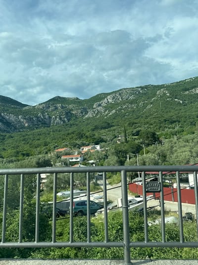 A mountain covered in vegetation is viewed through a metal railing. A small village with houses is located on the mountain's slope. The sky is cloudy. The location is Kaluđerac, Montenegro. Parked cars and boats are visible behind the railing.