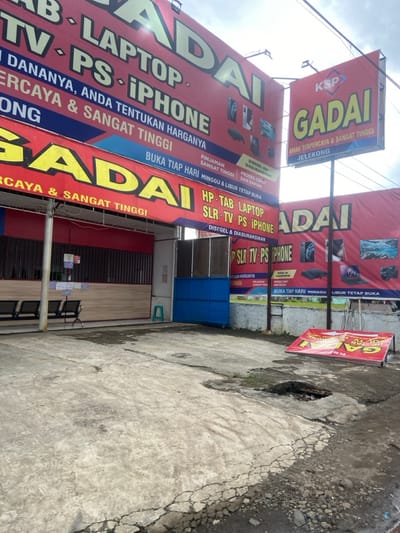 The image shows a pawn shop in Baleendah, Indonesia, located at latitude -7.021 and longitude 107.657. The shop's facade is covered in red and white signage advertising pawn services for electronics, including laptops, TVs, PlayStations, and iPhones. The shop is open, with a blue gate and an open door, and there are four black metal benches lining the wall in front of the shop. The concrete ground in front of the shop is cracked and uneven, with a small hole near the bottom right corner of the image.  A blue metal gate is partially open in the background, and an upside-down sign for the pawn shop is lying on the ground. The sky is overcast and gray. The image was taken on a weekday.