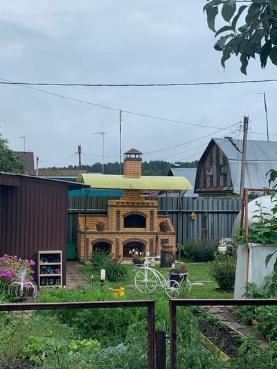 The image depicts a backyard scene, likely in Dubnik, Russia, on an overcast day. In the center, a brick barbecue oven with a yellow canopy is prominent. To its left is a dark brown wooden shed. A white metal decorative bicycle with a planter is positioned in front of the barbecue. Various plants and flowers, including pink and purple blooms, are visible throughout the yard. A metal fence runs across the foreground, separating the viewer from the garden. In the background, several houses with corrugated metal roofs and a greenhouse are visible, along with utility poles and electrical wires. The ground is covered in green grass and a brick pathway.