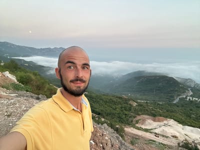 A man in a yellow polo shirt is taking a selfie on a rocky hillside overlooking a misty valley. The man has a shaved head and a beard. In the background, mountains are shrouded in fog, and a crescent moon is visible in the pale sky. The valley below is green and appears to be a mix of natural landscape and some construction or development, with piles of earth and what looks like construction vehicles. A winding road is visible in the distance. The scene appears to be located in or near the village of Lapčići, Montenegro, given the characteristic mountainous terrain and coastal proximity.