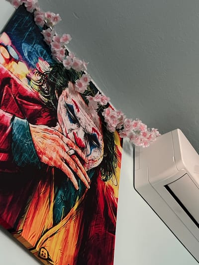 The image shows a close-up of a painting of the Joker character, with its face partially obscured by hands. The painting is adorned with a string of artificial pink cherry blossoms that drape across the top left corner and extend towards the right. To the right of the painting, a white air conditioning unit is mounted on a light gray wall. The painting's vibrant colors and the delicate flowers create a stark contrast with the functional appliance. The scene is indoors, with the wall and ceiling suggesting a domestic or commercial interior. The location in Debelt, Bulgaria, is not visually discernible from the image itself.