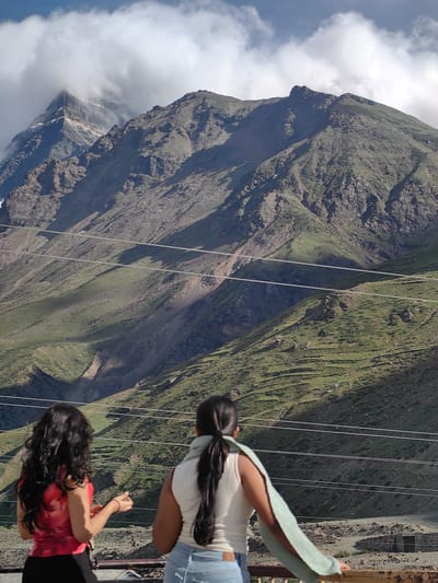 The image captures a scenic mountain landscape with two individuals standing in the foreground, looking out at the view. The mountains are covered in lush green vegetation with rocky outcrops and deep ravines. Clouds are present in the sky, partially obscuring some of the mountain peaks. Multiple power lines run horizontally across the frame, adding a man-made element to the natural scene.
In the foreground, from left to right, a person with long, dark, curly hair wearing a red and black outfit stands with their back to the camera. They appear to be holding something in their hands and are looking towards the mountains. To their right, another person with dark hair pulled back in a ponytail is wearing a white top and blue jeans. They have a light green scarf draped over their shoulders and are also facing away from the camera, observing the mountain vista. The environment suggests a location in Yamling, India, known for its mountainous terrain.