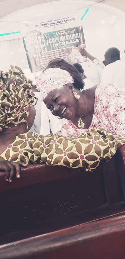 The image depicts an indoor scene, likely a church hall, in Kaduna South, Nigeria. A banner in the background, partially obscured, advertises an event hosted by "ST. JOHN'S ANGLICAN CHURCH, MAKERA, KADUNA SOUTH." The banner also mentions "Harvest of SUPERNATURAL INCREASE" and "2023 JUVENILE YOUTH HARVEST."
In the foreground, two individuals are partially visible. The person on the right, a woman with dark skin, is wearing a pink lace headwrap and a pink floral dress. She is adorned with large, gold-toned earrings and a matching pendant necklace. She is looking downwards and laughing, with her mouth open. Her left arm is visible resting on what appears to be a wooden bench.
The person on the left is wearing a headwrap with a brown and yellow floral pattern. Only their upper body and head are visible from the side. Their left hand is resting on the wooden bench in front of them. Both individuals are positioned as if seated on a bench, with the bench being a prominent element in the lower part of the image. The lighting suggests an interior setting, possibly with artificial light.