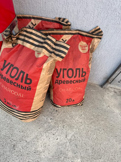 Two red paper bags filled with charcoal are lying on a textured gray concrete surface next to a rough, light-colored wall. The bags have black and beige striped bands running vertically. The top bag displays "УГОЛЬ Древесный" in large black Cyrillic letters, with "CHARCOAL" and "20 A" below it. A circular red and white label on this bag reads "Экологически Чистый Продукт!" (Ecologically Pure Product!). The bottom bag is partially visible and shows similar text, including "УГОЛЬ Древесный CHARCOAL" and "5 кг ±7%". The setting appears to be outdoors, possibly near a building entrance, given the wall and the concrete. The lighting suggests daytime.