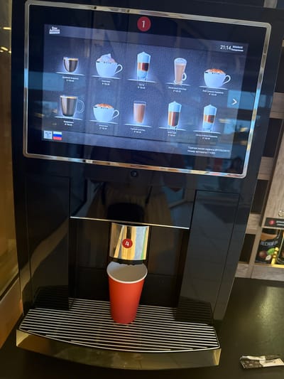 The media file shows a close-up, slightly angled, exterior view of a modern, black coffee machine. The machine has a large touchscreen display on its left side, showing a menu of coffee drinks with images and prices. Visible text on the screen includes "21:14", "2023/04/22", "Понедельник", and various coffee names like "Капучино", "Латте", "Моккачино", "Американо", and "Двойной Эспрессо", along with prices in Rubles (₽). The brand "DON ANTONIO ESPRESSO" is also visible on the screen.
To the right of the screen, a red paper cup is positioned under a spout, suggesting that coffee is being dispensed or has just been dispensed. Above the cup, there's a metallic dispenser area with a red circular button containing the number "4". The background shows a slightly out-of-focus interior scene, possibly a cafe or break room, with shelves containing some products, including what appears to be coffee-related items. One of these products has Cyrillic text, one of which appears to be "ПЕСНОЙ". A small white packet with text and graphics is hanging on the wall to the right. The overall lighting suggests it is daytime indoors. The scene is devoid of people.