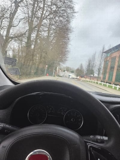 The image displays a forward-facing view from the interior of a vehicle on an overcast day. A black, textured steering wheel with a central circular red emblem occupies the lower half of the frame. Behind the steering wheel, the vehicle's instrument cluster is partially visible, showing two circular gauges and various indicator lights. Control stalks extend from the steering column on both the left and right sides. The windshield, visible above the dashboard, is covered with numerous small water droplets, indicating damp conditions. Two rectangular stickers are affixed to the lower left portion of the windshield.
Outside the vehicle, a wet, paved two-lane road stretches into the distance. On the left side, a dense aggregation of tall deciduous trees, bare of leaves, lines the road, suggesting winter or early spring. A red traffic cone is positioned near the roadside on the left. Further down, several light-colored buildings are visible amongst the trees. On the right side of the road, a low, white, segmented barrier or fence runs parallel to the roadway. Beyond this barrier, a modern building with brick cladding, dark windows, and a dark roof structure stands. In the distance, a white utility van is visible on the right side of the road. The sky is uniformly grey and overcast. This scene depicts a road environment within the city of Braine-l'Alleud, Belgium. The specific street or address within Braine-l'Alleud cannot be determined from the image.