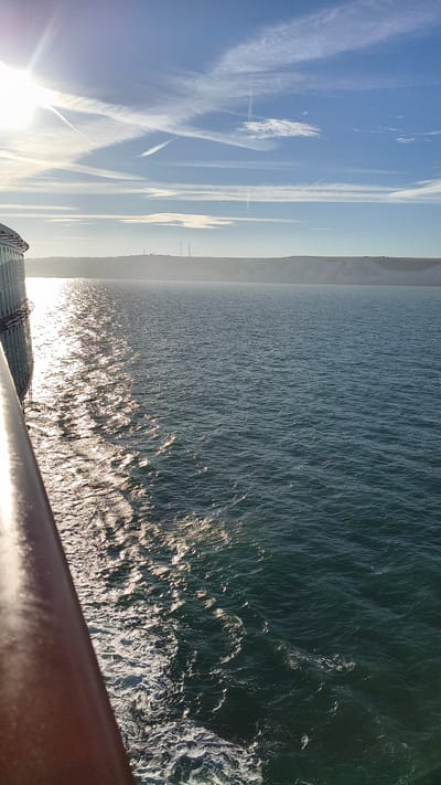 The image captures a view from a vessel moving on the sea, likely a ferry, approaching the coast of Dover, United Kingdom. The sun is high and bright in the sky, casting a strong reflection on the choppy water. The sea is a deep blue-green, with white foam marking the wake of the vessel on the left side of the frame. In the background, a low, chalky cliff face is visible, characteristic of the White Cliffs of Dover. Several indistinct structures, possibly towers or industrial buildings, are silhouetted against the horizon on top of the cliffs. The sky is a clear blue, with thin, streaky cirrus clouds, some of which appear to be contrails from aircraft. The railing of the vessel is visible in the foreground on the left, and part of the vessel's exterior, a light-colored structure with vertical lines, is also present on the far left. The overall impression is one of a bright, clear day at sea with land in sight.