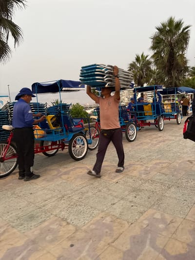 A man in a t-shirt and dark pants walks to the right, carrying a stack of folding chairs on his head. Another man in a blue shirt, hat, and black pants stands at the front of a row of blue four-wheeled pedal vehicles. Palm trees are visible in the background. The scene appears to be near the beach in Ancón, Peru.
