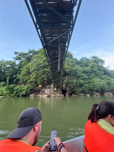This is a photograph taken from a boat on a body of water, looking up at a large, wooden and metal bridge. The bridge appears to be an elevated walkway or pedestrian bridge that spans over the water and a vegetated cliff face. On the cliff face, there are several torches burning, casting an orange glow. A person in light-colored clothing is visible standing on a walkway near the torches. The water is green, and the surrounding environment is lush with dense green trees and foliage. In the foreground, two people are visible from behind, also on the boat. One person is wearing a black baseball cap and an orange life vest, and is holding a smartphone. The other person is wearing an orange life vest and has their hair in a ponytail. The sky above is a clear blue. The scene is likely located near the city of Yên Sơn Ward, Vietnam, given the architectural style of the bridge and the surrounding natural environment.