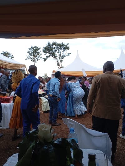 The image captures an outdoor social gathering in Shimbi Mashariki, Tanzania, under a large canopy with gold and white draped fabric overhead, extending to white pointed tents in the background. Numerous adults are present, mingling and engaging in activities that suggest a celebration, possibly including dancing, as indicated by the movement of a woman in a patterned light blue dress in the midground.
In the foreground, the backs of several people are visible, including a man in a blue suit, a woman with curly hair in a mustard-yellow dress, and a man in a brown jacket on the right. A table with a white tablecloth is partially visible in the bottom right, adorned with a white rose centerpiece in a vase, a plastic water bottle, and glass bottles. To the left, a table with a red patterned box, possibly a gift, is present.
The ground appears to be bare earth. In the midground, an older woman is seated on a chair under a decorated canopy, suggesting she might be an honored guest. The sky is overcast, indicating a daytime event, likely late afternoon, with soft, diffused lighting. Scattered trees are visible beyond the tents in the background. The atmosphere appears lively and convivial, with people relaxed and interacting. No discernible text is visible in the image.